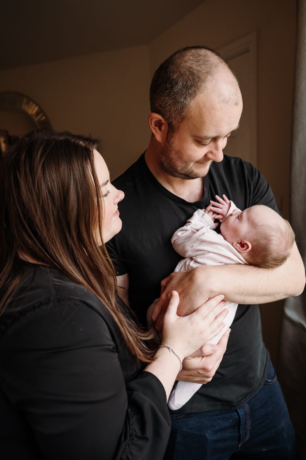 Dad is holding his new daughter and wife is looking at them by Natural newborn photographer