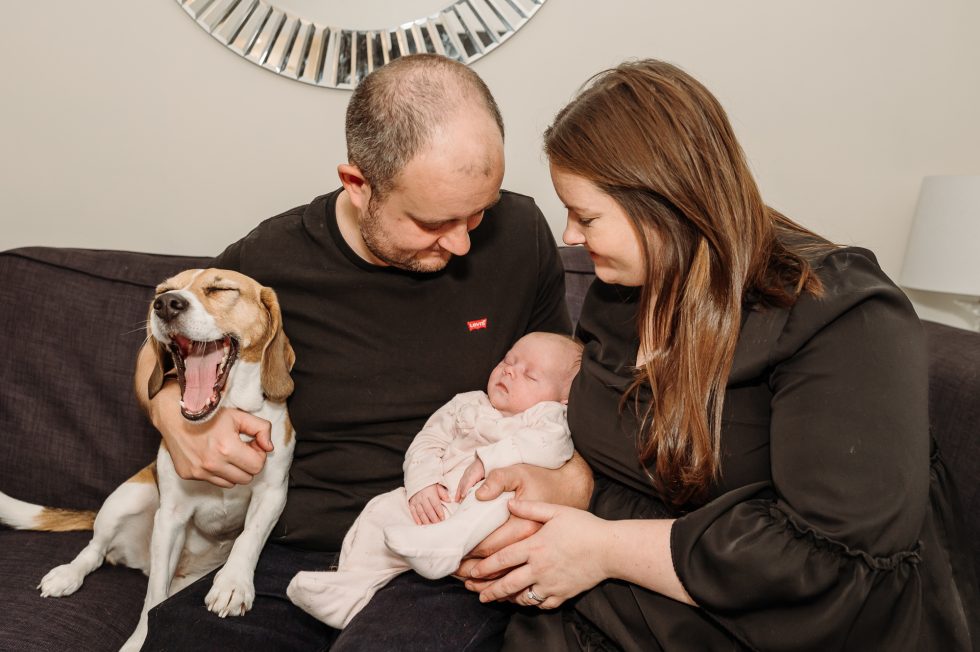 Mum and dad are holding their newborn girl on the sofa and the family dog is sat with them by Natural newborn photographer