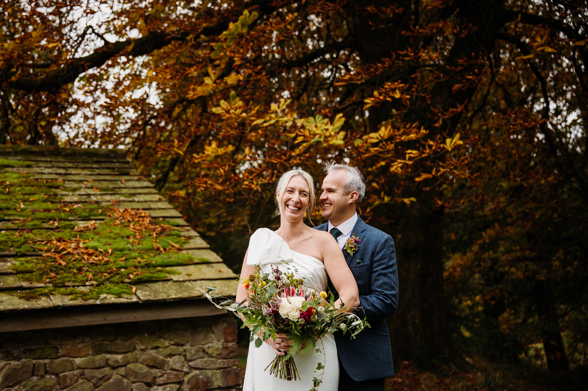 Groom is hugging his new wife and there are gorgeous autumnal colours on the trees behind them by Browsholme Hall Wedding Photographer