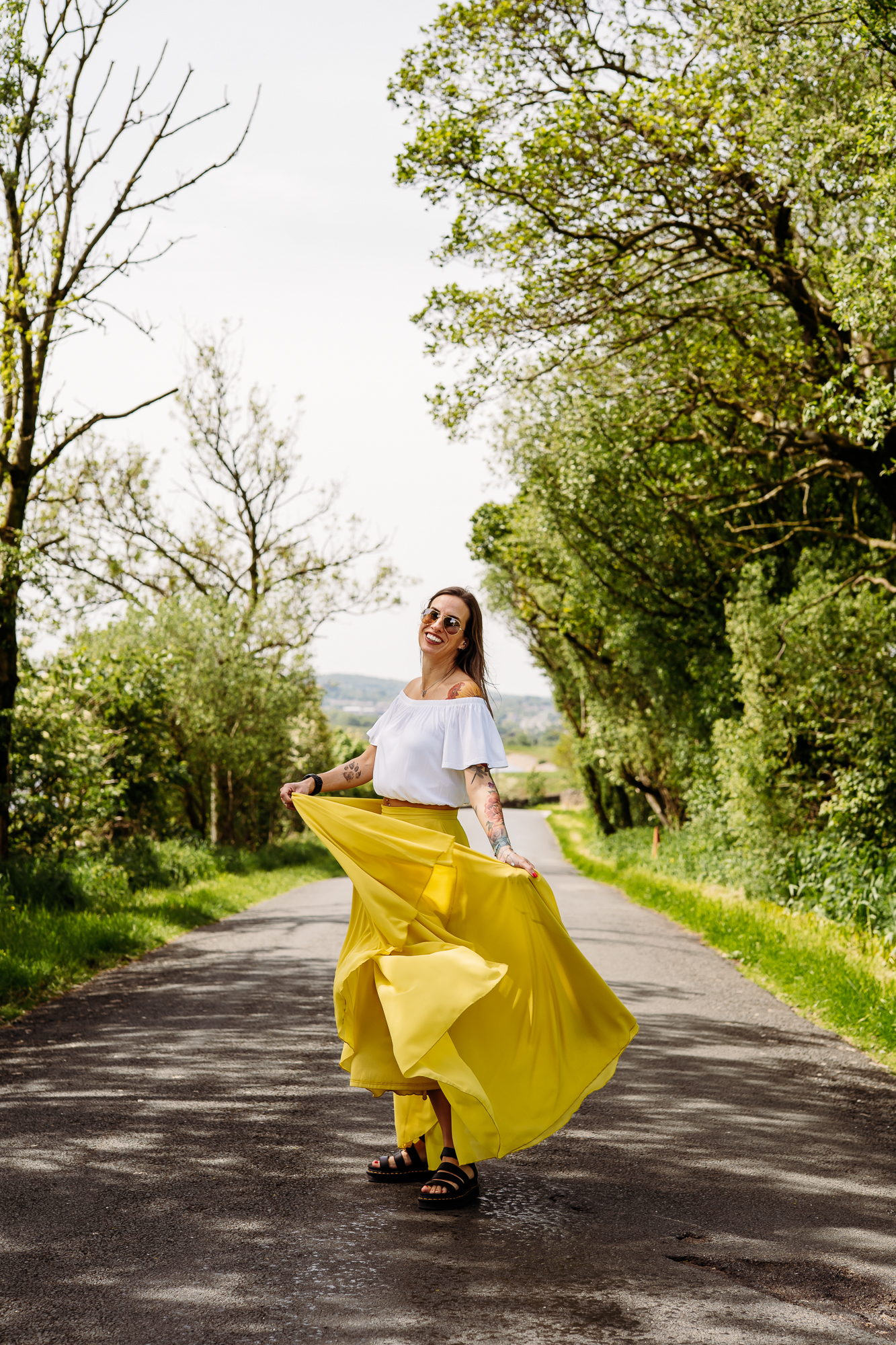 Lancashire wedding photographer, Jane Green is stood in the middle of a county road in the Ribble Valley and swishing her long yellow skirt around and laughing