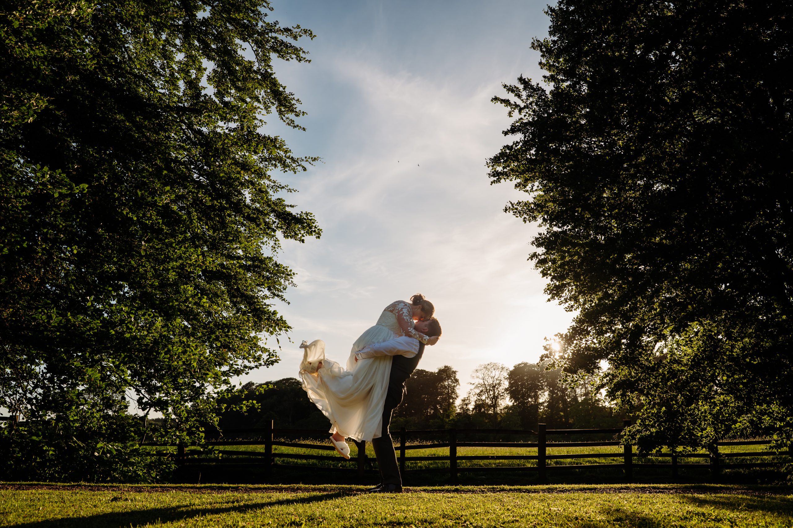 Groom is lifting his new wife up in the garden. There is a beautiful sunset in the background and they are framed by trees by Mitton Hall wedding photographer