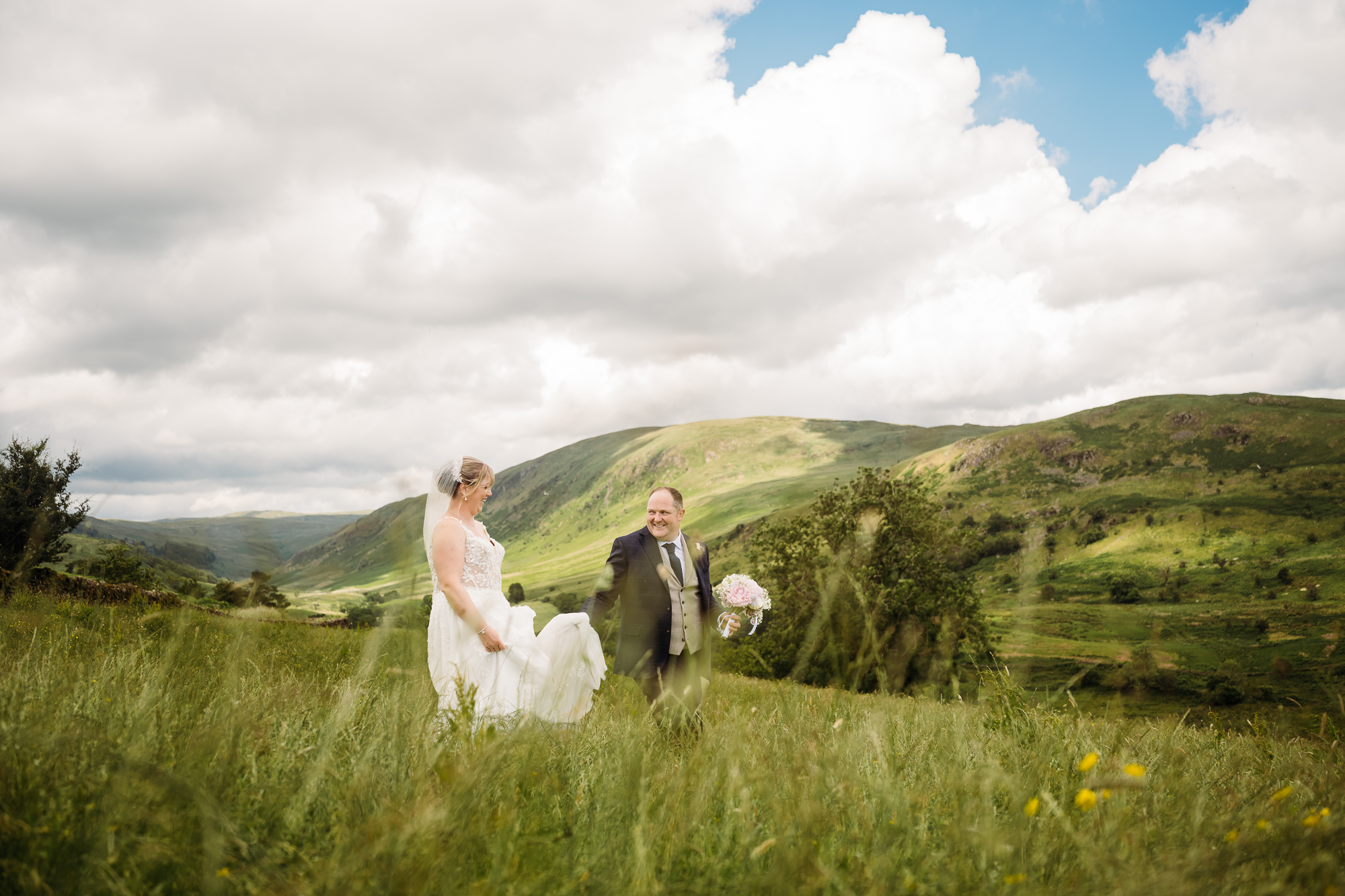 Bride and groom walking through a meadow with a Cumbrian valley behind them